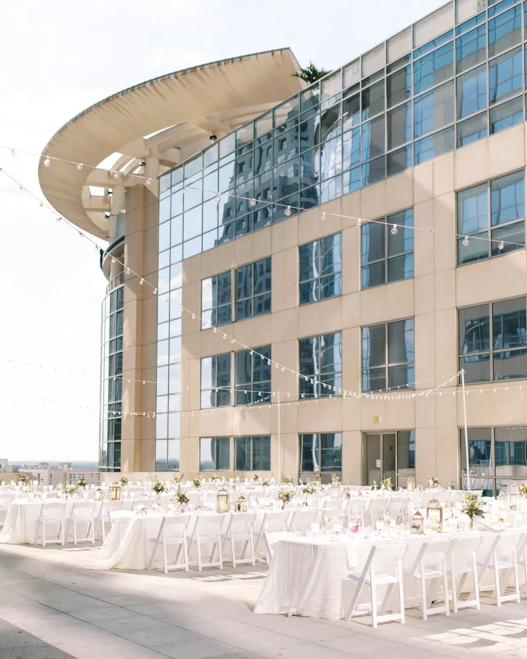 rooftop central florida venue the balcony orlando1 - Wedding Venues Collections Elegant outdoor rooftop wedding reception setup with white folding chairs, white linen-draped banquet tables, gold lantern centerpieces, and string lights against a modern glass building facade at The Balcony Orlando in Orlando, Central Florida