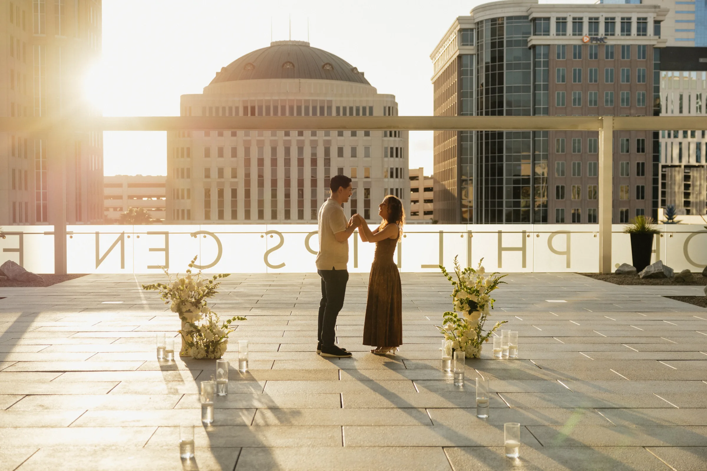 Couple dancing during a romantic rooftop proposal at the Dr. Phillips Center in Orlando Florida surrounded by white floral arrangements and candles at golden hour, photographed by Vanessa Oppedal Photography