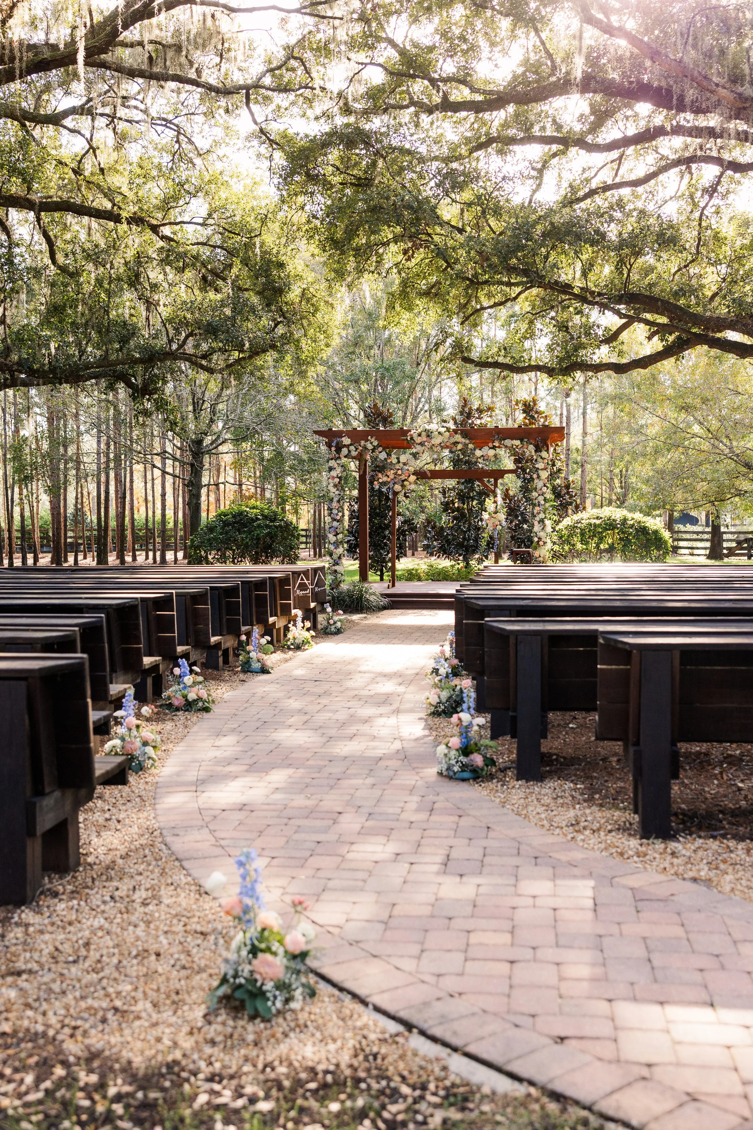 rustic central florida wedding venue club lake 1 - Wedding Venues Collections Outdoor wedding ceremony at Club Lake wedding venue in Central Florida featuring dark wood pew benches lining a brick aisle path leading to a wooden arbor adorned with white and blush floral arrangements, set beneath sprawling live oak trees draped with Spanish moss