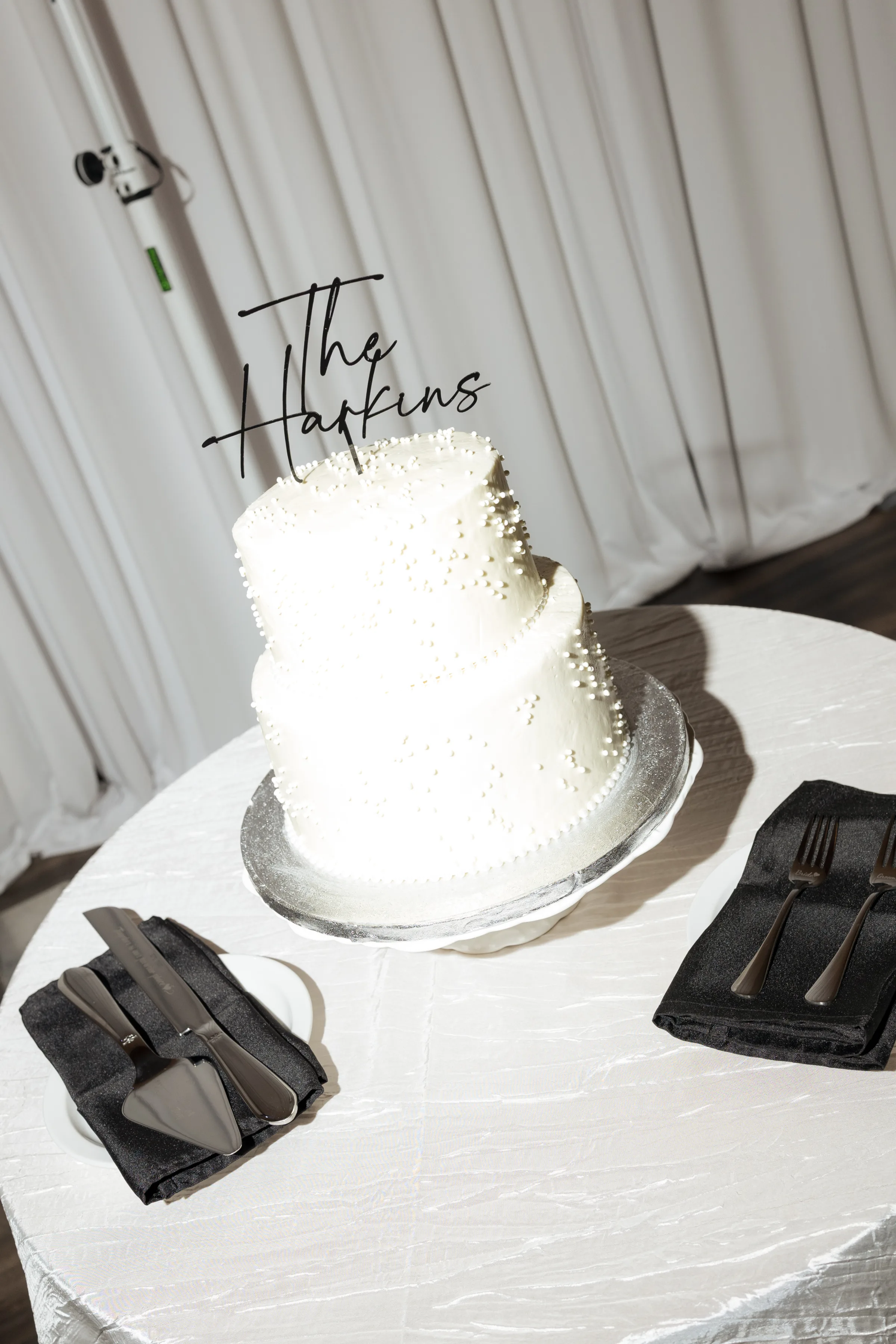 Elegant two-tier white pearl-studded wedding cake with The Harkins cake topper displayed on a silver stand with black napkins and silverware at an Orlando Central Florida wedding reception