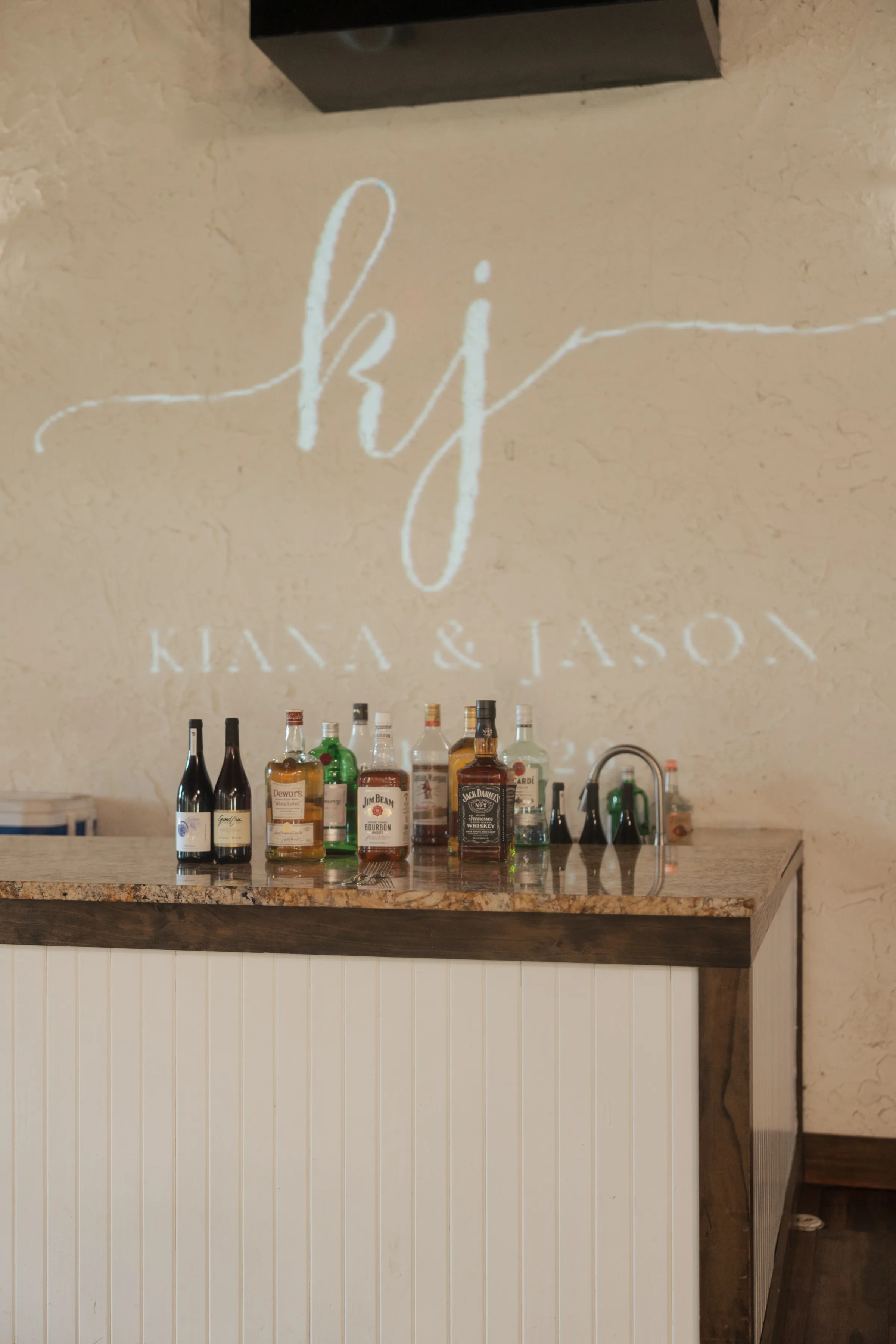 Elegant wedding bar setup with liquor bottles including Jack Daniels and Jim Beam on a granite countertop beneath a projected monogram reading Kiana and Jason at a Central Florida wedding venue