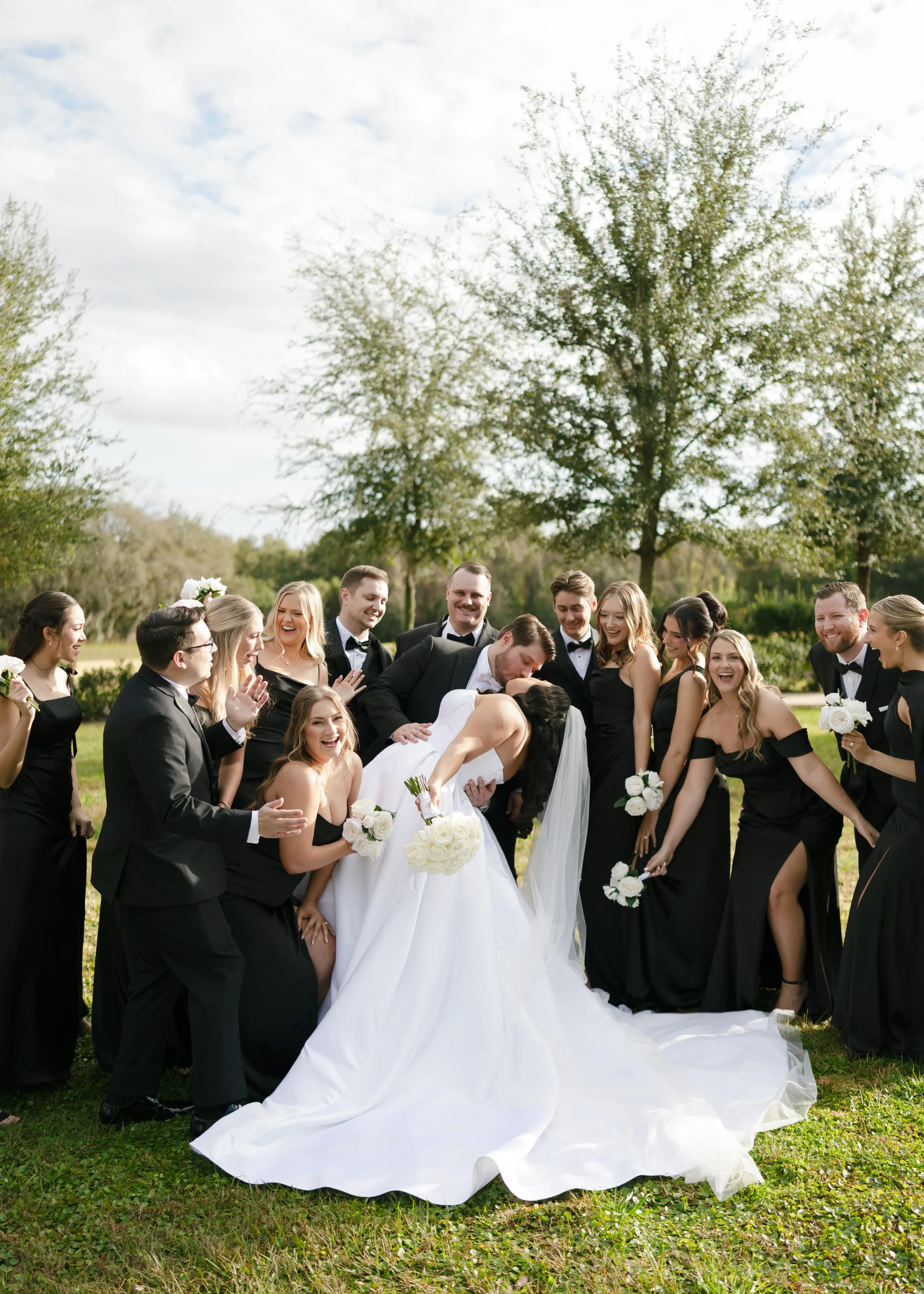 Bride and groom share a romantic dip kiss surrounded by their cheering wedding party dressed in black formal attire at an outdoor Central Florida wedding venue