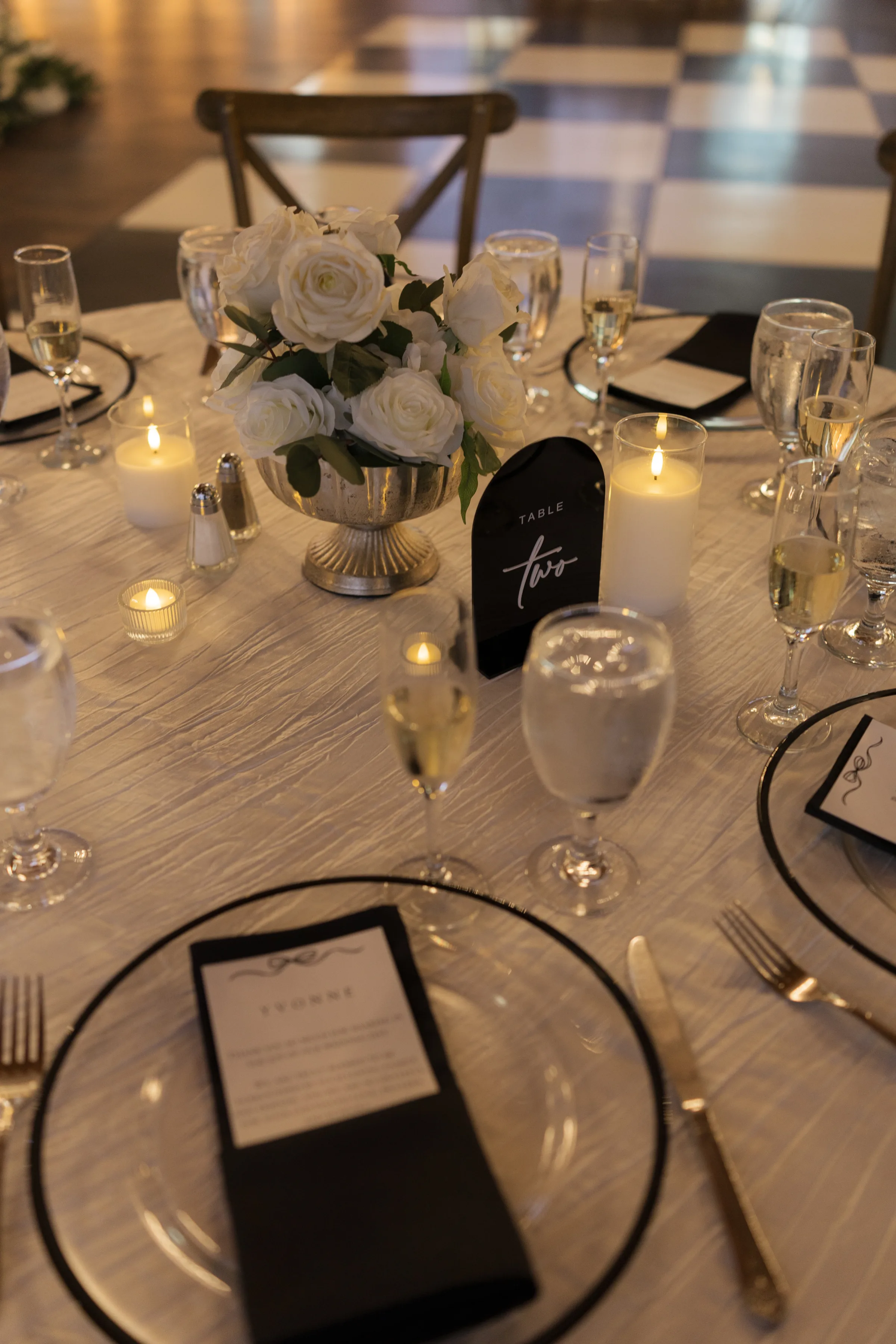 Timeless black and white wedding reception table setting with white rose centerpiece in silver compote, black-rimmed charger plates, gold flatware, lit candles, and a black acrylic Table Two sign on a wood-grain linen in Orlando, Florida