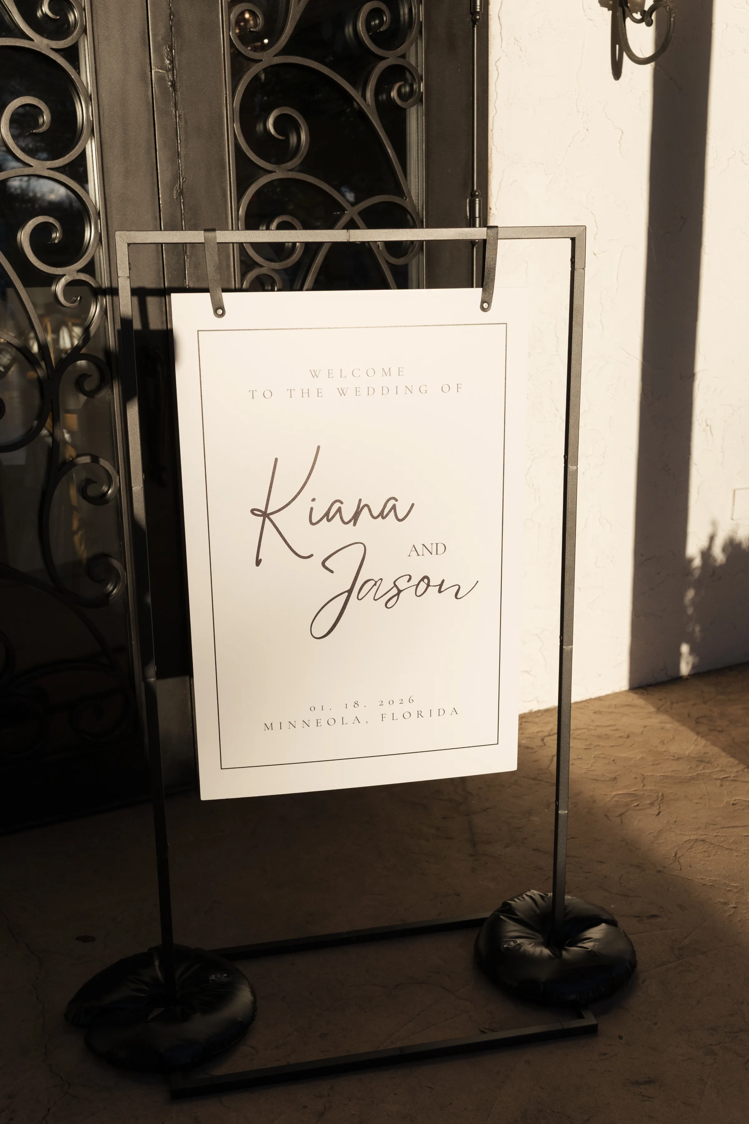 Elegant black and white wedding welcome sign for Kiana and Jason displayed on a modern iron stand outside a venue in Minneola, Florida, dated January 18, 2026
