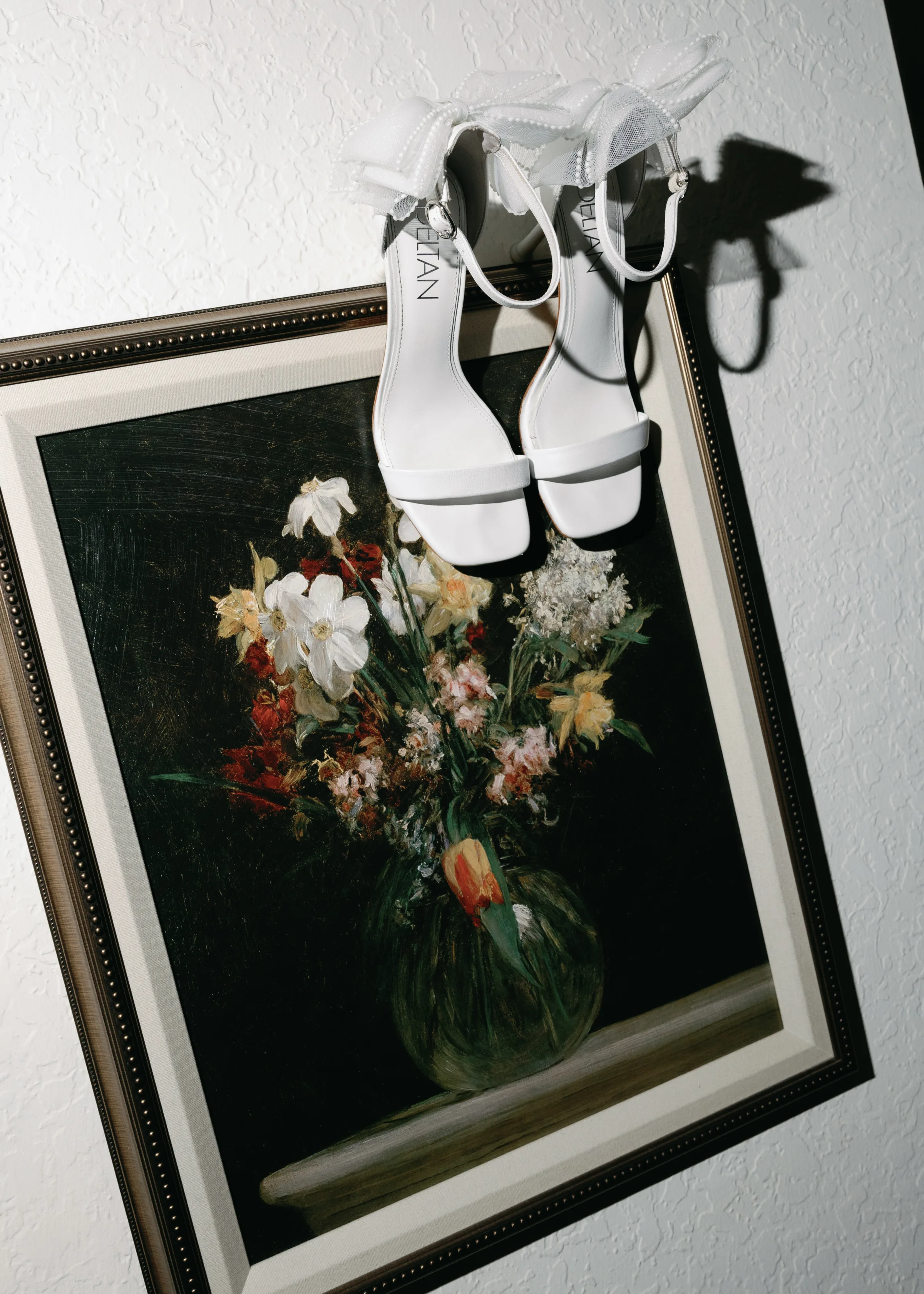 White strappy bridal heels by Celian hanging over a dark classical floral oil painting, styled for a wedding detail shot in Orlando, Florida